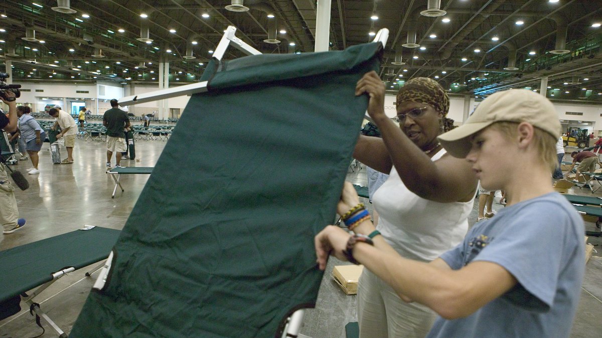 Volunteers setting up rows of cots inside a large shelter hall