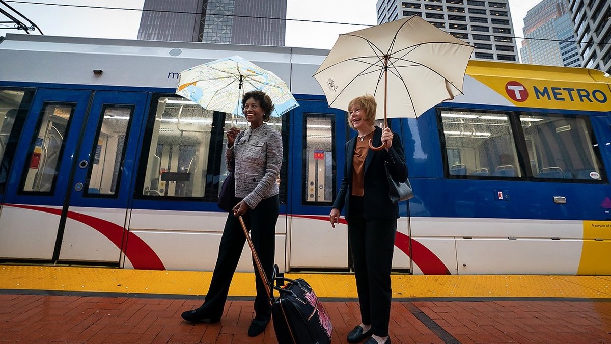 Two riders with umbrellas standing in front of a modern light rail train at a city station